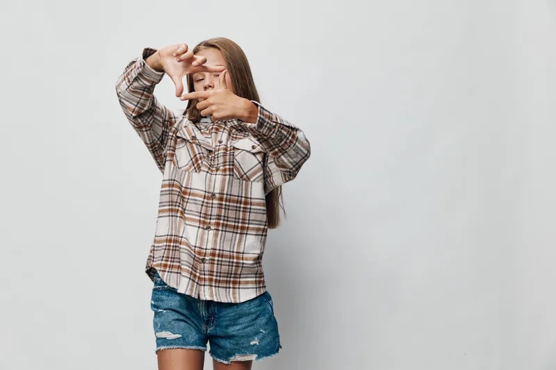 Girl in plaid shirt and denim shorts posing with hands framing face on white background for creative photography or expression of art and youth style.
