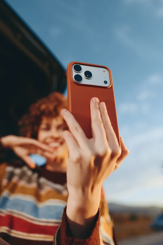Woman lifestyle selfie outdoors, closeup hands holding red smartphone, blue sky background, blurred friend in striped top, casual travel in daylight, relaxed moment