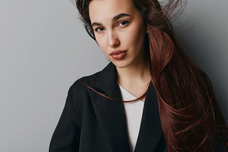 Young woman with long dark hair and natural makeup wearing black coat and white top posing against gray background looking confident and stylish with subtle expression in studio portrait.