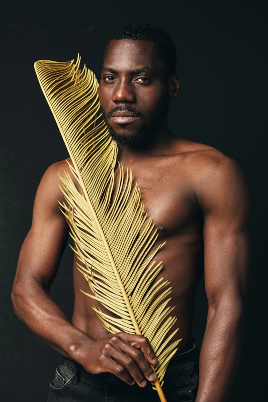 African man with dark skin holding large golden palm leaf in creative conceptual visual style. Shirtless male model posing with artistic expression on black background.