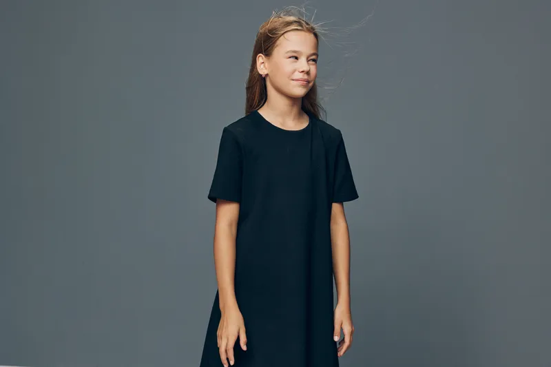 Girl posing in black dress with hands by sides looking slightly away on gray background. Child with long hair, casual outfit, standing and calm expression in studio portrait.