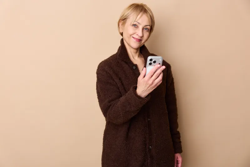 Woman portrait, holding smartphone, wearing coat, beige studio background, smiling, relaxed communication, modern lifestyle, adult, close up, vertical frame