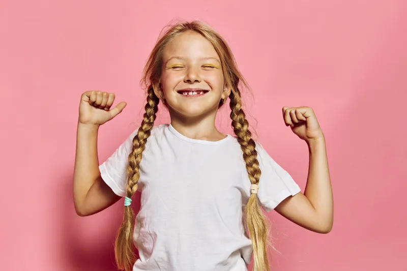 Happy girl with braided hair smiles showing missing teeth while flexing arms on pink background.