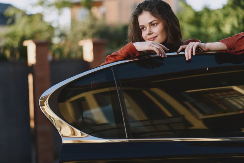 A smiling woman leans on a car door in a sunlit outdoors setting, capturing a casual lifestyle moment. A candid portrait of everyday confidence and modern urban living