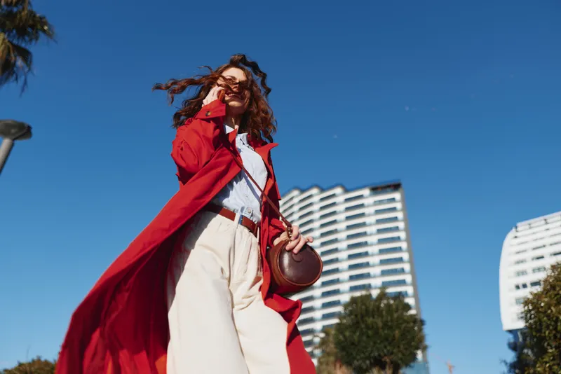 A stylish young woman with curly hair strolls outdoors in a red trench coat over a white shirt and beige trousers, posing confidently amid modern high rises.