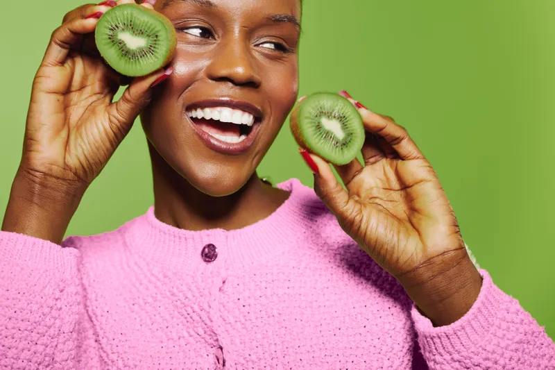 Happy African woman with dark skin holds two halves of kiwi fruit near face, smiling brightly. Concept of healthy lifestyle, fresh fruit, wellness, and natural beauty on green background.