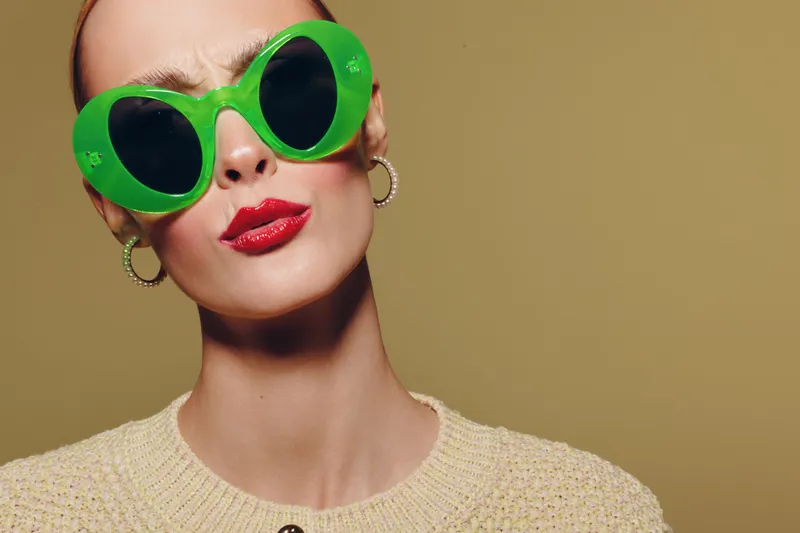 Stylish woman with short hair wearing large green sunglasses and red lipstick poses against beige background. Fashion portrait with bold accessories and confident expression.