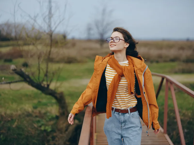 Woman lifestyle outdoors, smile with glasses, walking on a wooden bridge, orange jacket and scarf, striped shirt, calm daylight, nature background, leisure moment