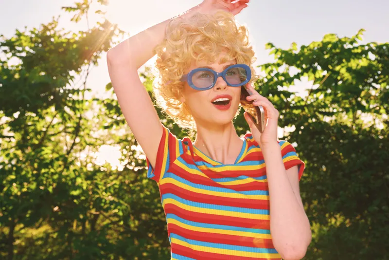 Young woman with retro style outdoors, striped colorful shirt, round blue sunglasses, curly hair, joyful expression, sunny day, candid lifestyle shot with warm film color and cheerful mood, isolated
