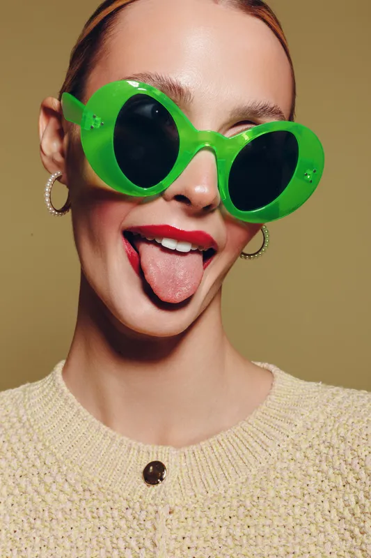 Young woman with green sunglasses and red lipstick sticks out tongue. Close-up portrait on beige background. Concept of fun, fashion, and playful attitude in studio lighting.