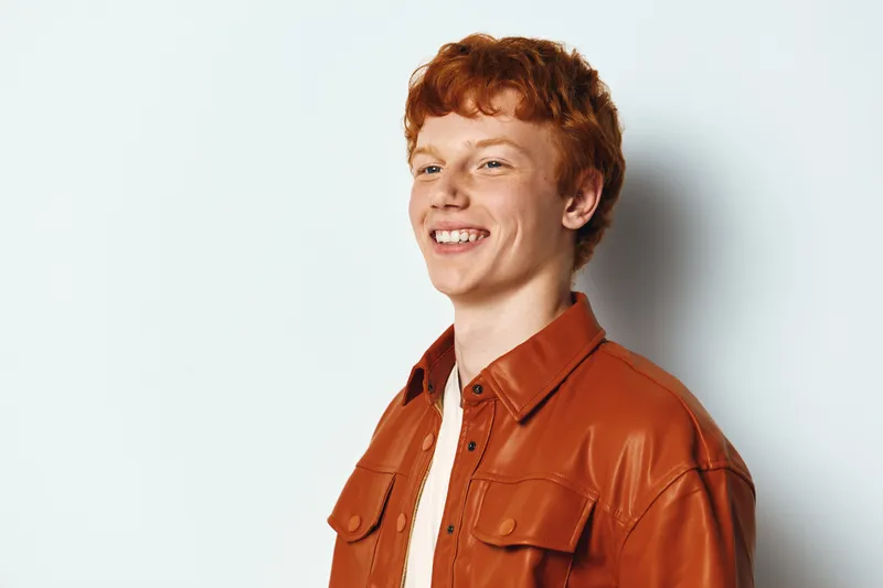 Young man with red hair smiling and wearing a brown jacket standing against white background. Casual style with confidence and natural expression in studio.