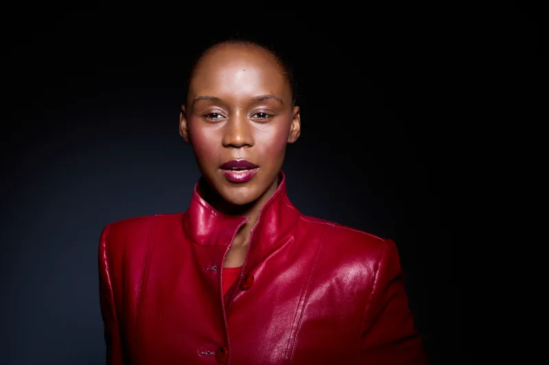 Portrait of confident black woman with dark skin wearing a red leather jacket. Studio lighting highlights her smooth skin and strong facial features on a dark background. Fashion and beauty concept.