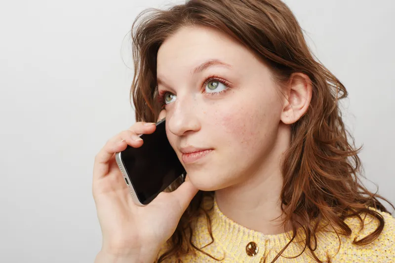 Young woman with curly hair wearing yellow sweater talks on smartphone, looking away thoughtfully. Concept of communication, technology, and casual lifestyle in indoor setting.