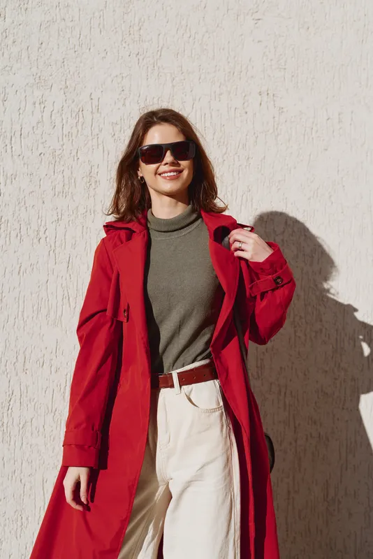 Young woman with long brunette hair wearing sunglasses and a red coat smiles confidently while standing against a textured beige wall in bright sunlight.