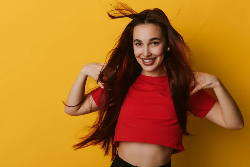Young woman with long hair wearing red crop top smiling and posing with hands near shoulders on yellow background. Happy female model with casual style and natural makeup.