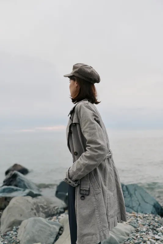 Woman in grey coat and hat stands on rocky shore looking at calm sea under overcast sky. Concept of solitude, contemplation, and autumn outdoor fashion by the water.