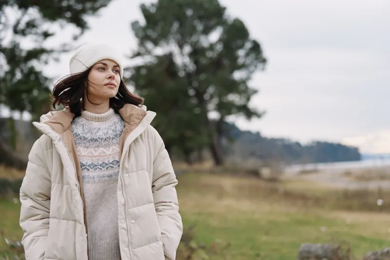 Woman in a beige puffer coat and knit sweater stands outdoors by a windy coastline, calm expression, trees in background, candid fashion moment in nature.