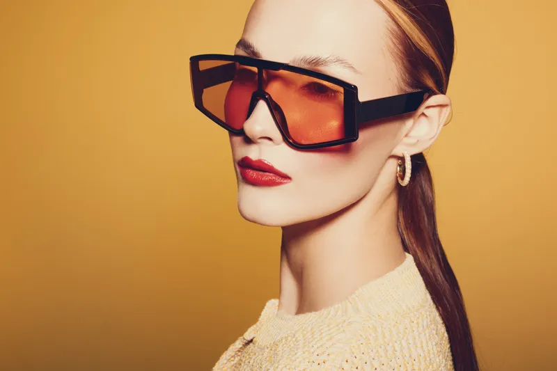 Stylish woman with large orange sunglasses and red lipstick poses against a warm yellow background. Fashion portrait with modern accessories and confident expression.