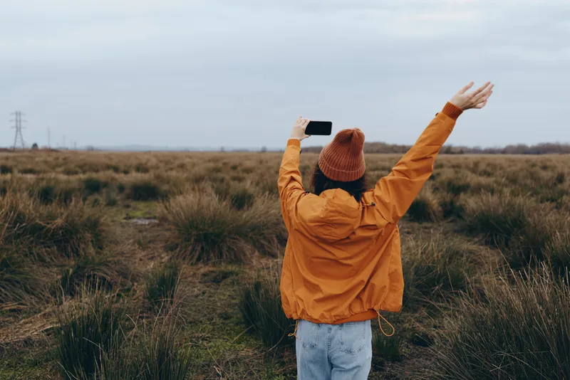 Woman lifestyle outdoors in a wide field, wearing orange jacket and beanie, holding smartphone near her chest, arms raised in wind, looking up, joyful portrait, calm spring nature mood