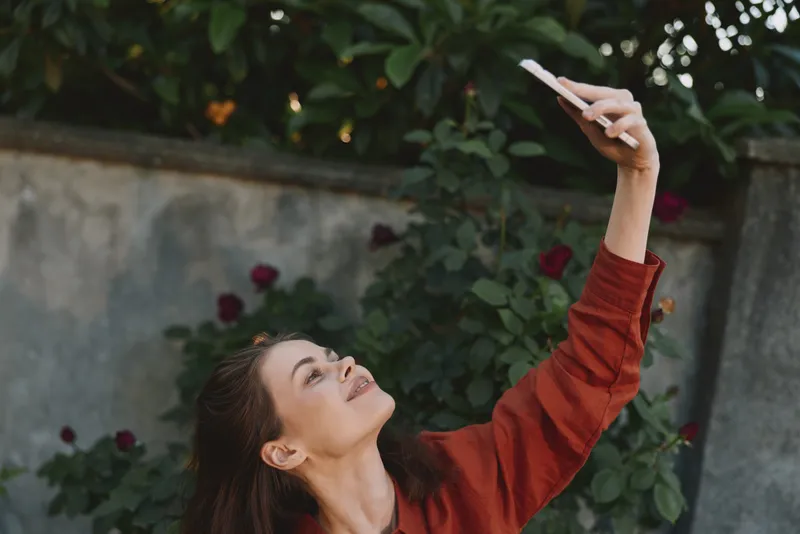 Woman takes a casual selfie outdoors in a garden with roses, wearing a red sweater, enjoying a relaxed lifestyle moment. bright moment captured against sunlight