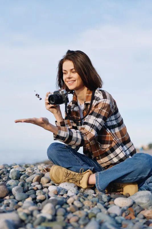 Photographer woman kneels on a rocky beach, holding a camera and capturing a candid moment outdoors. Casual plaid shirt, jeans, boots, sunny sky and calm sea backdrop.