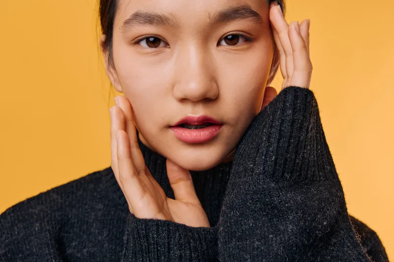 Young woman with natural makeup wearing dark sweater posing with hands near face, looking at camera on bright orange background for fashion and beauty portrait.