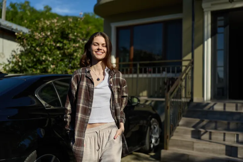 A smiling woman stands in front of a house on a sunny day, dressed in casual layers. A candid lifestyle moment highlighting everyday confidence and relaxed fashion