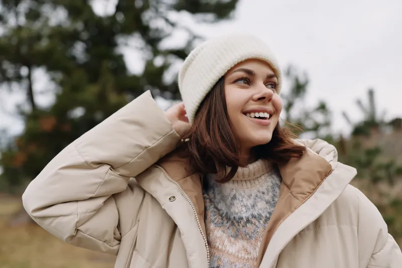 Woman in a beige jacket and knit sweater outdoors, smiling in a beanie, relaxed pose against a natural background, lifestyle fashion shot capturing warmth, comfort and seasonal style
