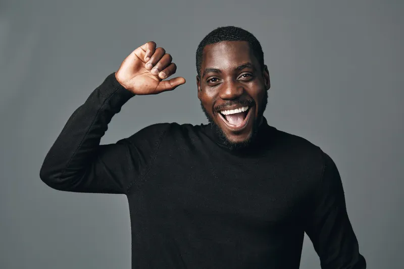 African American man with dark skin wearing black sweater smiling and raising hand in creative visual designer style studio portrait on gray background. Happy male model.