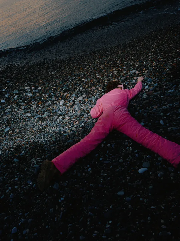 Pink pants worn by a lone figure lie motionless on a pebble beach beside dark water at dusk, creating a moody scene of stillness, texture and contrast in outdoor coastal light