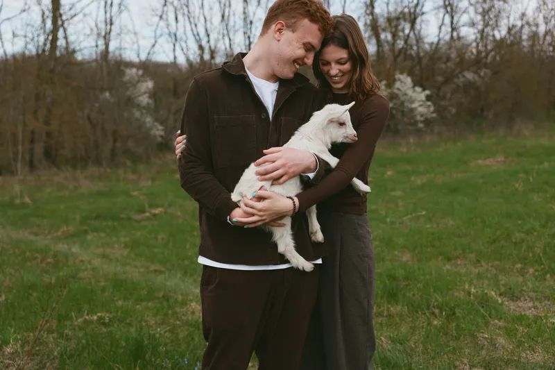 Happy couple outdoors with a playful goat kid, they embrace softly in a grassy field, warm casual outfits, peaceful countryside mood, lifestyle moment of affection and animal companion