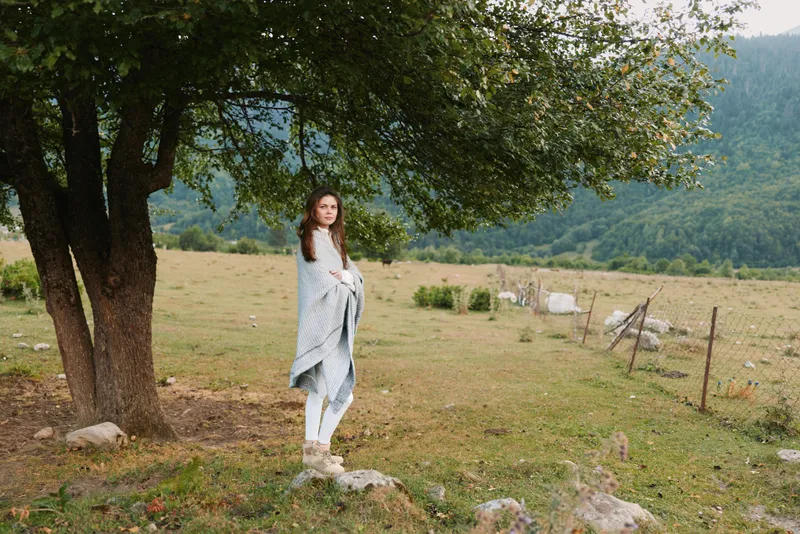 Young woman in casual clothes stands under tree in rural landscape with mountains in background, wrapped in blanket, enjoying peaceful countryside lifestyle.