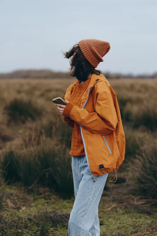 Woman lifestyle outdoors in autumn, standing in a grassy field, wearing an orange coat, knit hat and scarf, walking forward and looking down, calm mood, rural countryside background