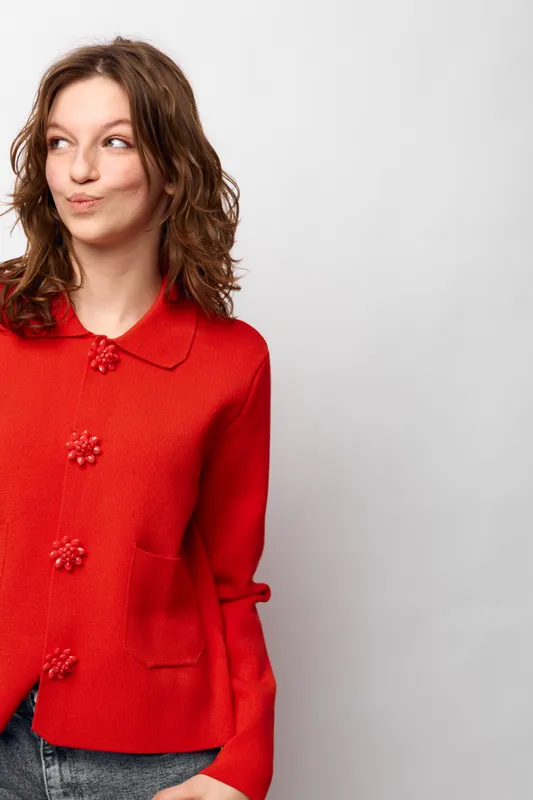 Young woman in red sweater looks sideways with playful expression. Casual style, curly hair, and light background create a relaxed and cheerful mood.