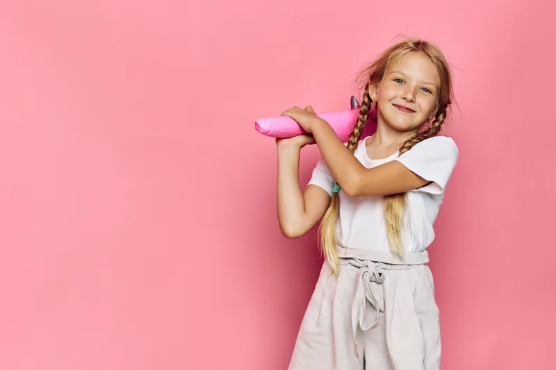 Happy girl with blonde hair holds a pink hairbrush while smiling on a bright pink background.