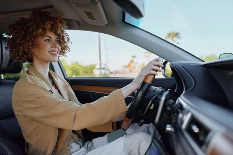 Smiling woman wearing a beige jacket driving a car, side profile, bright daylight on a street. Lifestyle scene highlighting travel, confidence, and everyday moments in a sunny setting.