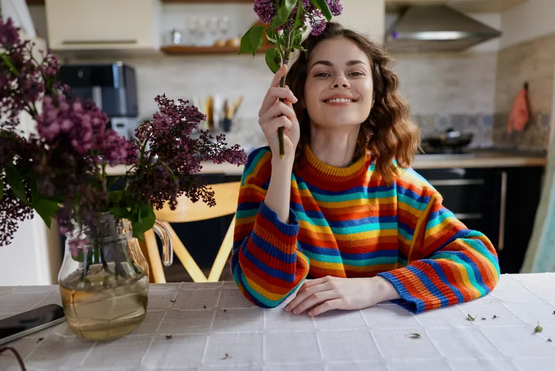 A smiling woman sits at a kitchen table in a colorful striped sweater, holding a bouquet. Natural light fills a bright home scene, with flowers and casual still life, creating a warm, inviting mood.