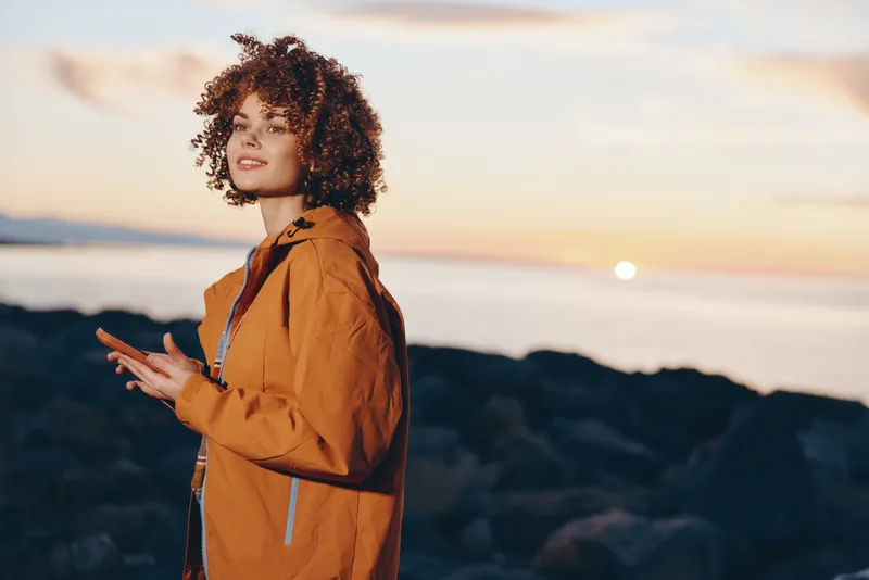 Woman lifestyle portrait outdoors at sunset on the coast near the sea, orange jacket, curly hair, smiling and looking into the distance, wind and warm light, cinematic golden hour
