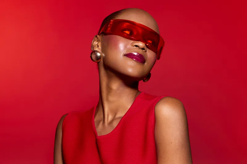 Elegant black woman with dark skin wearing red sleeveless top and red visor sunglasses poses confidently against red background. Concept of fashion, style, and beauty.