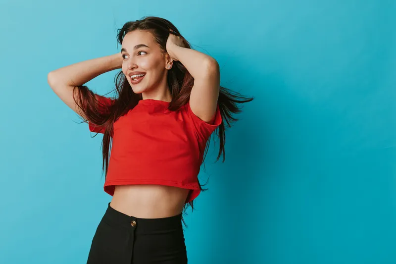 Young woman in red crop top and black pants posing with hands in hair against blue background. Casual style, smiling face, long hair and confident expression in studio.