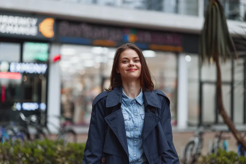Young woman with long brown hair smiles confidently while standing outdoors wearing a dark trench coat and denim shirt in an urban environment.