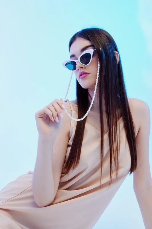 Fashionable woman with long hair wearing stylish sunglasses and a beige dress poses against a light blue background. Concept of beauty, fashion, and modern style.