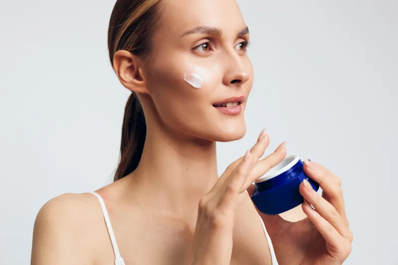 Young woman applying face cream with white moisturizer, holding blue cosmetic jar, beauty skincare wellness self care concept, close up portrait, light studio background, soft studio lighting,