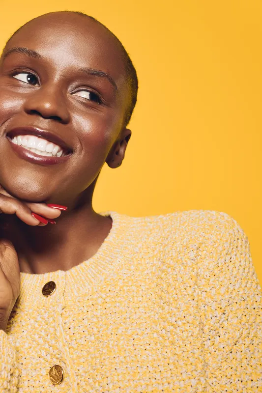 Smiling black woman with dark skin wearing a yellow knitted sweater poses against a bright yellow background. Concept of happiness, fashion, and confident female portrait.