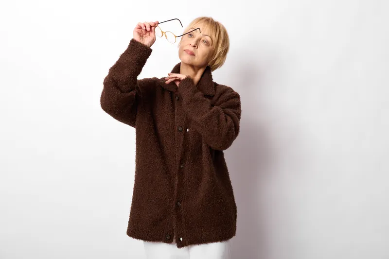 Woman with glasses adjusts eyewear in studio portrait, blonde adult wearing brown coat, playful confident expression, indoor background, vision and eye