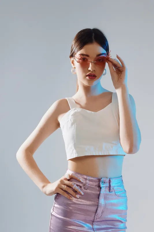 Young woman in a white crop top and shiny pants adjusts pink sunglasses. Studio portrait with soft lighting on a neutral background. Concept of woman beauty fashion.