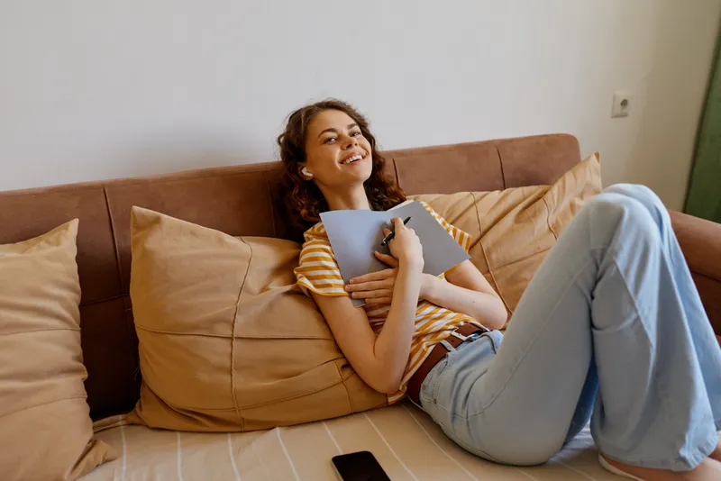 Young woman relaxes on a home sofa, smiling while hugging a notebook, wearing a striped tee and light jeans, creating a warm casual lifestyle moment.