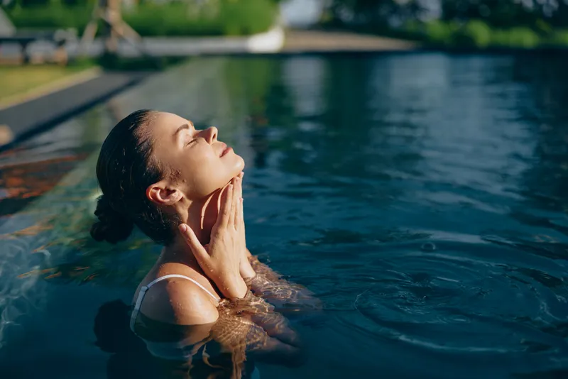 A woman relaxes in a sunlit outdoor pool, head tilted back, hands at her neck, savoring warm water and stillness. A lifestyle concept of calm, renewal, and aquatic leisure.