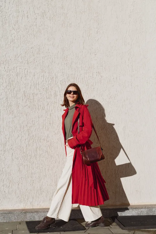 Young woman in a red trench coat and white pants walks confidently along a sunlit wall wearing sunglasses and carrying a brown leather shoulder bag on a clear day.