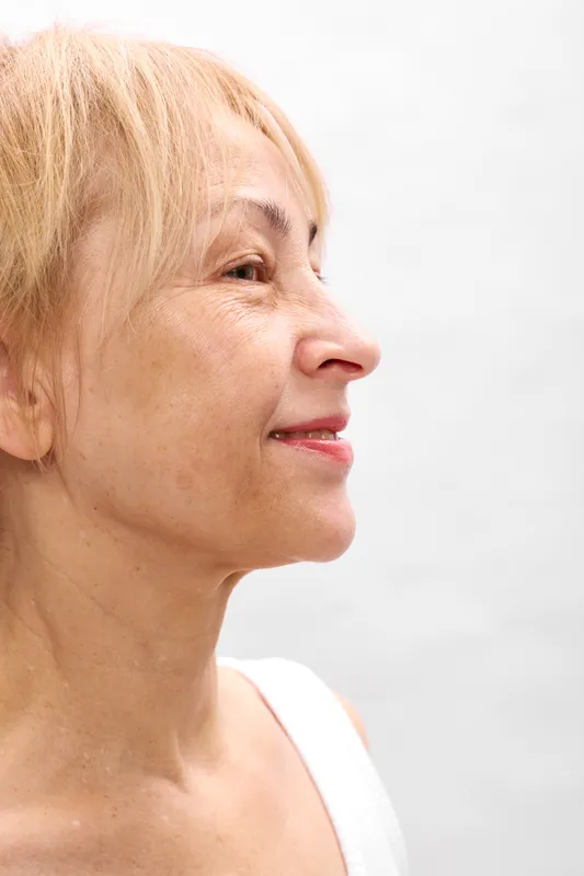 Elderly woman close up portrait profile, calm serene expression, short blond hair, soft skin texture, white background, beauty and aging, gentle skincare, copy space
